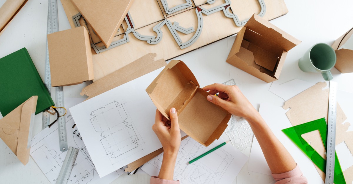 Packaging blueprints rest on a table with rulers and a pencil. Someone holds the actual constructed package.