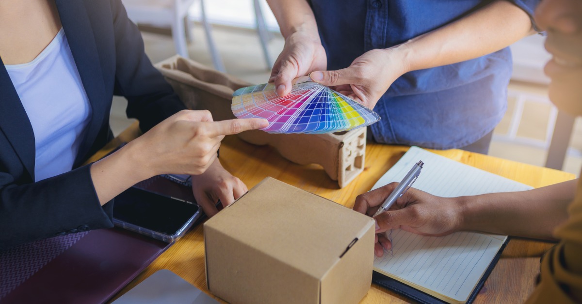 A group of people stands around a table with a packaging box in front of them. Someone fans out color swatches.