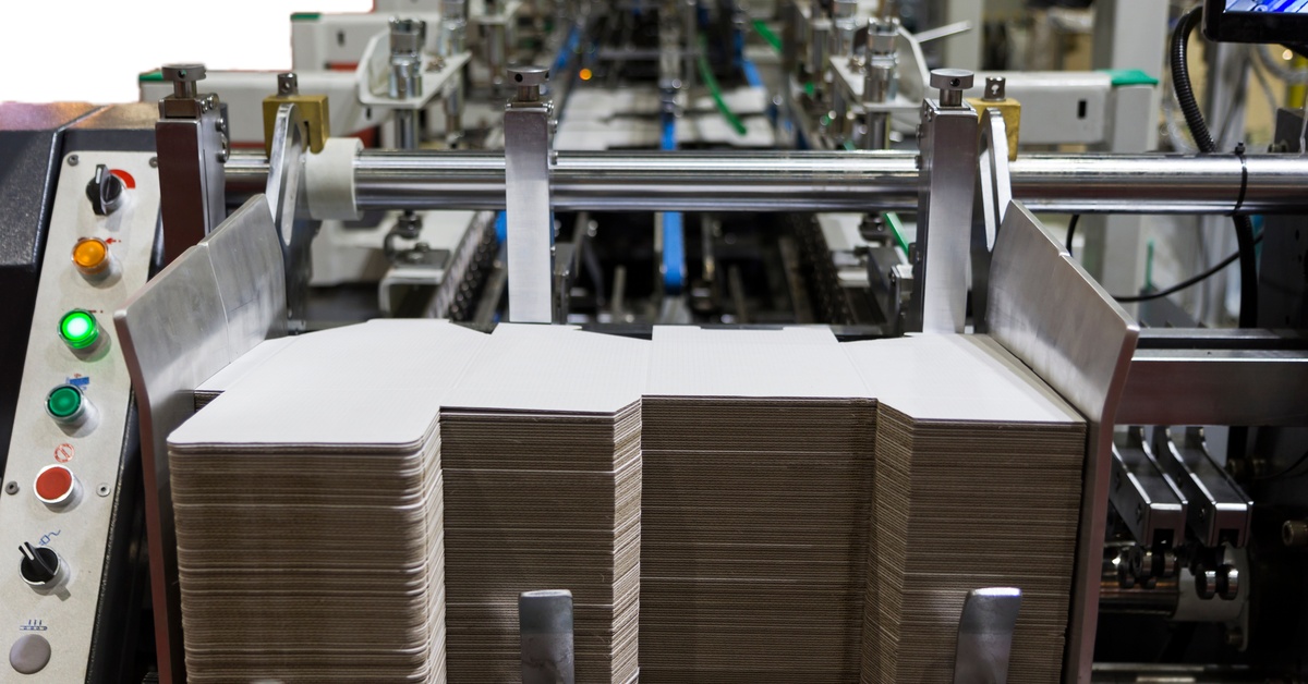 A large, neat stack of flat folding cartons sitting in a machine. A green button on the left panel is lit up.