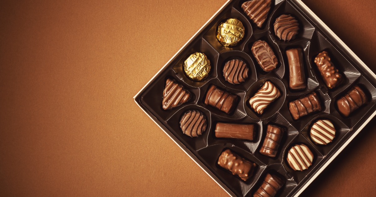 An assortment of chocolates neatly tucked into a tray within a box. The chocolates rest on a warm tan background.