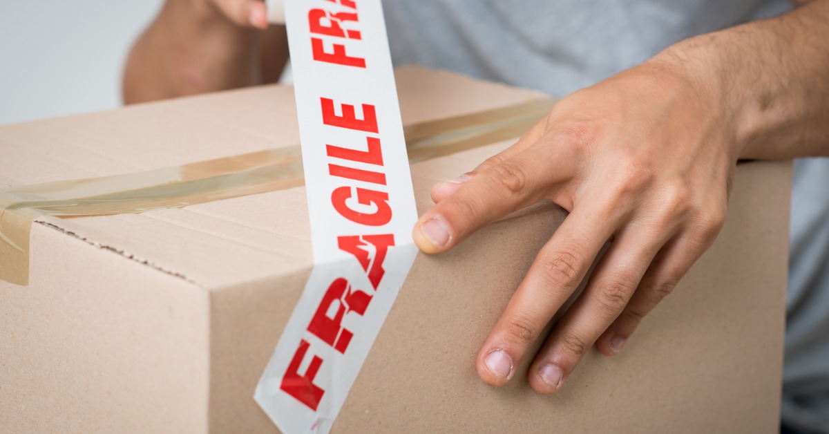 A close-up of someone applying white tape to the top of a cardboard box. The tape repeats the word