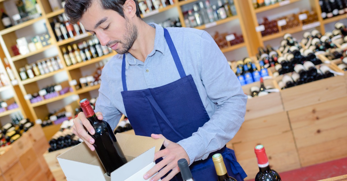 A man putting a wine bottle inside a cubic, white cardboard box placed on a wooden counter next to other bottles.