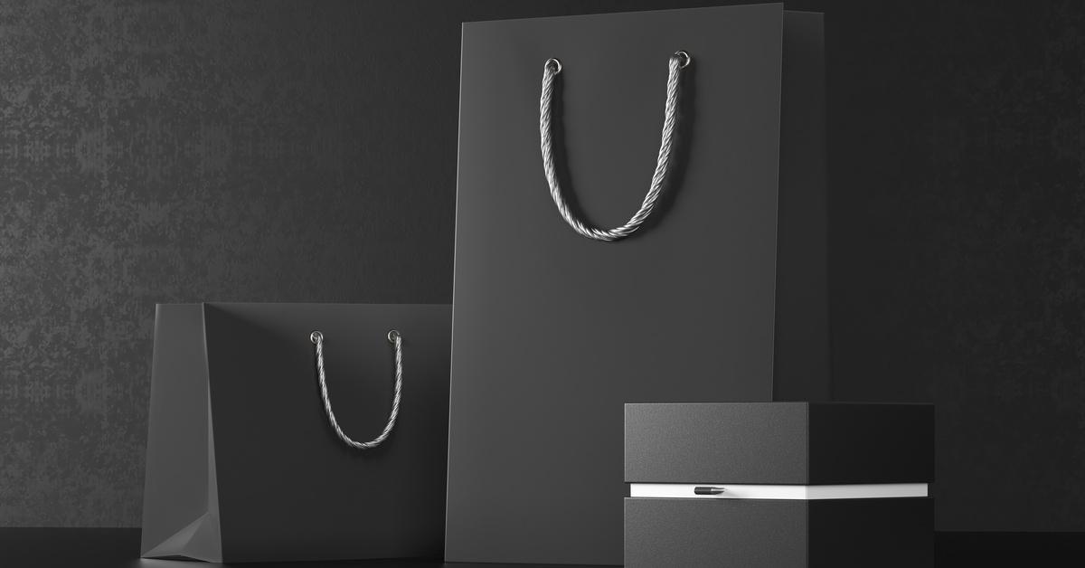 Two black paper bags in different sizes with grey braided handles against a black background. A small black box is next to the bags.