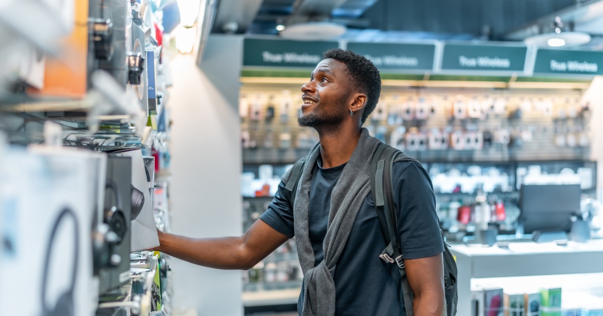 A man stands in a electronic store, looking at one of the product displays. He touches one item and looks up.