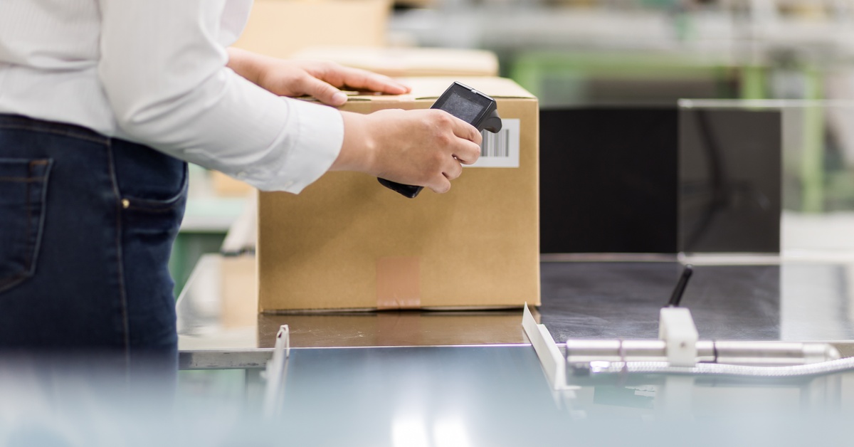 A person wearing a white shirt, using a scanner on a barcode placed on the side of a cardboard box in a warehouse.
