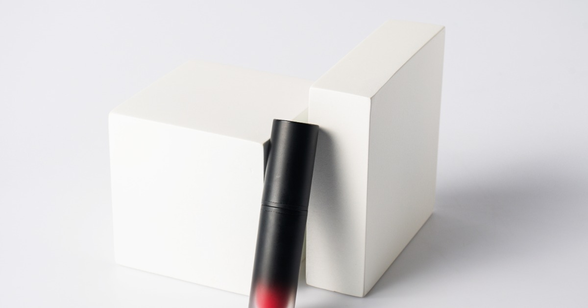 A close-up of a black lipstick container with a clear tip showing a red lipstick leaning against two cardboard boxes.