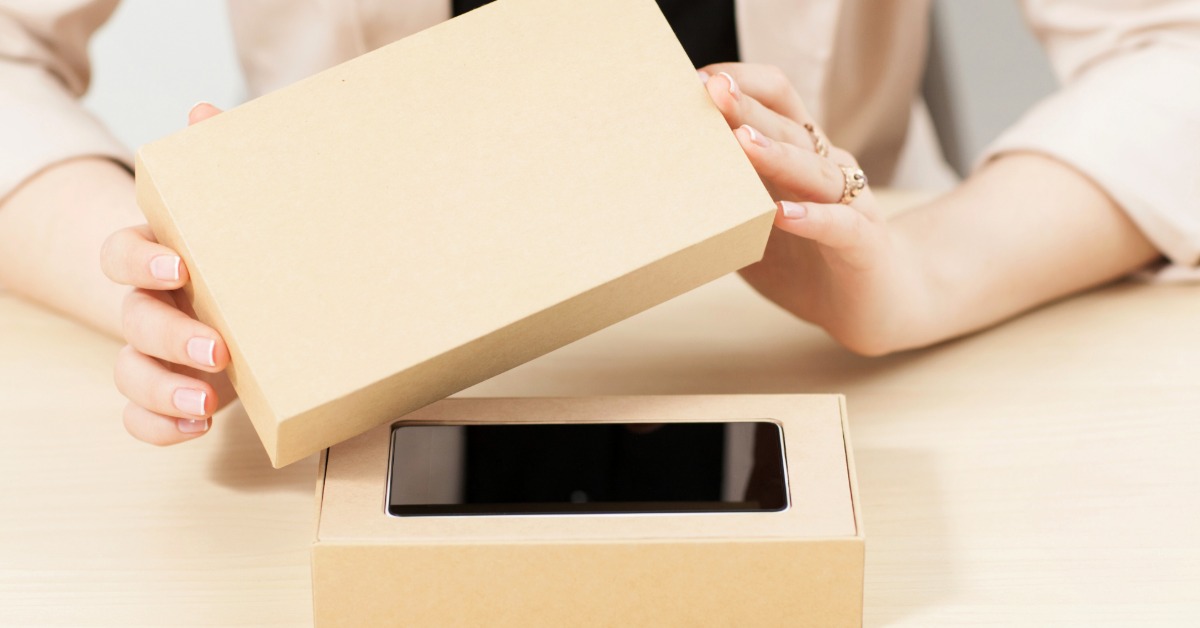 A close-up of two hands lifting the top of a brown box with a smartphone inside, placed on a yellow surface.