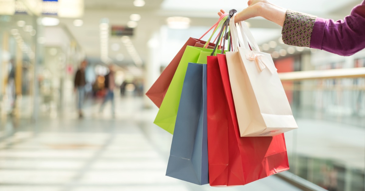 A close-up of a hand carrying multiple shopping bags of different sizes down a hall inside a large building.