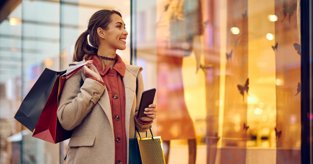 A woman wearing a brown coat walking next to a display window while carrying multiple shopping bags on her shoulder.